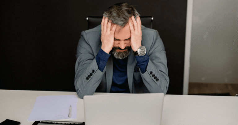 Hombre frustrado en su oficina apoyado sobre la mesa con las manos en la cabeza frente a un ordenador portátil, representando el impacto emocional y económico de una página web caída o hackeada por falta de actualizaciones de WordPress.